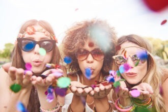 mulheres jovens e alegres soprando confetes e se divertindo juntas usando óculos de carnaval