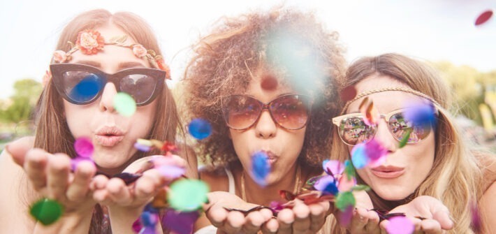 mulheres jovens e alegres soprando confetes e se divertindo juntas usando óculos de carnaval