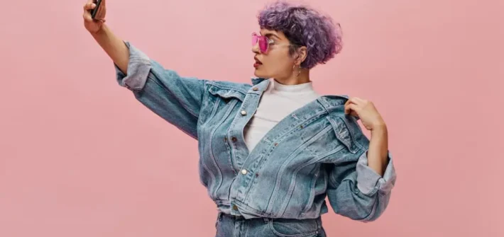 Mulher estilosa de jaqueta e calça jeans posando para selfie em fundo rosa claro