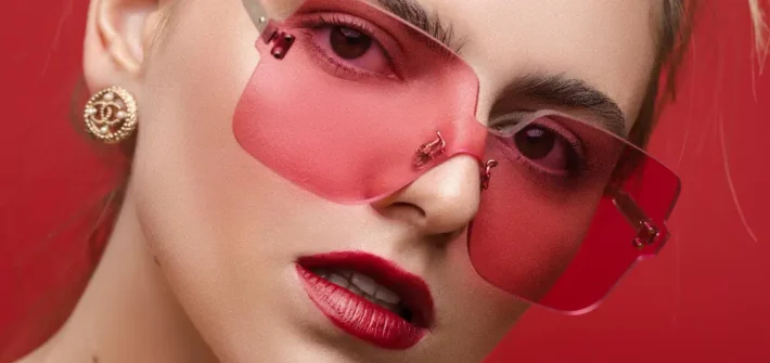 Mulher de maquiagem com óculos de lente rosa avermelhada em fundo vermelho