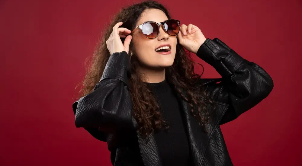 Mulher com roupas pretas ajustando óculos de sol no rosto enquanto sorri em fundo vermelho escuro