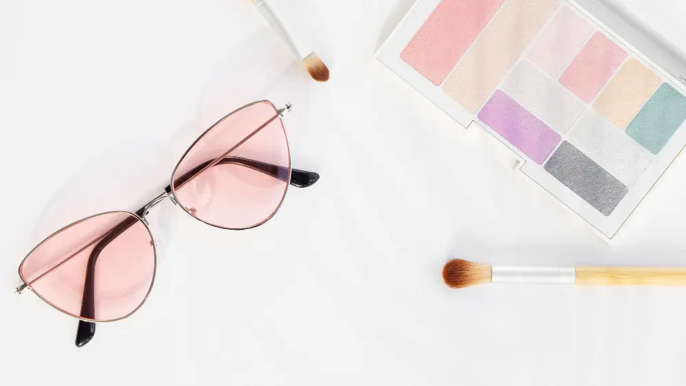 Armação de óculos com lentes rosa em cima de superfície branca com estojo de maquiagem e pincel 