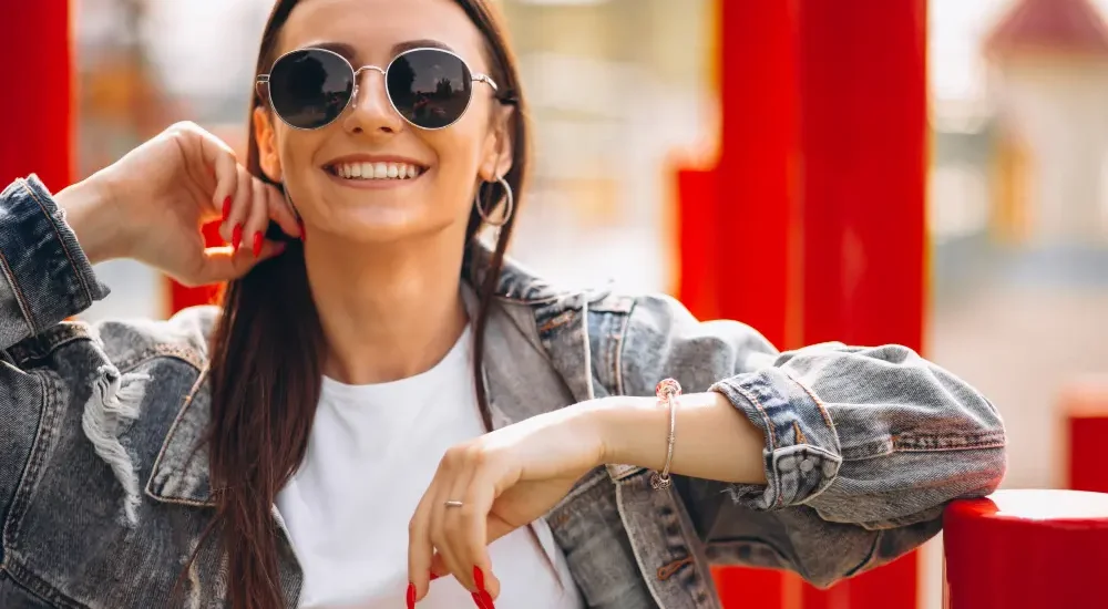 Mulher sorrindo usando combinação de óculos escuros e acessórios com jaqueta jeans, blusa branca e unhas pintadas de vermelhos