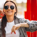 Mulher sorrindo usando combinação de óculos escuros e acessórios com jaqueta jeans, blusa branca e unhas pintadas de vermelhos