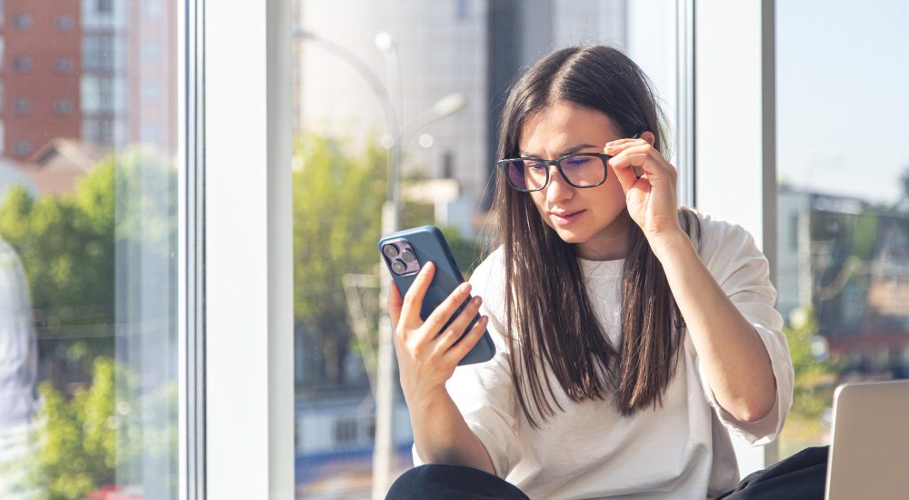 Mulher sentada ajustando os óculos enquanto olha para o celular forçando a visão