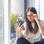 Mulher sentada ajustando os óculos enquanto olha para o celular forçando a visão