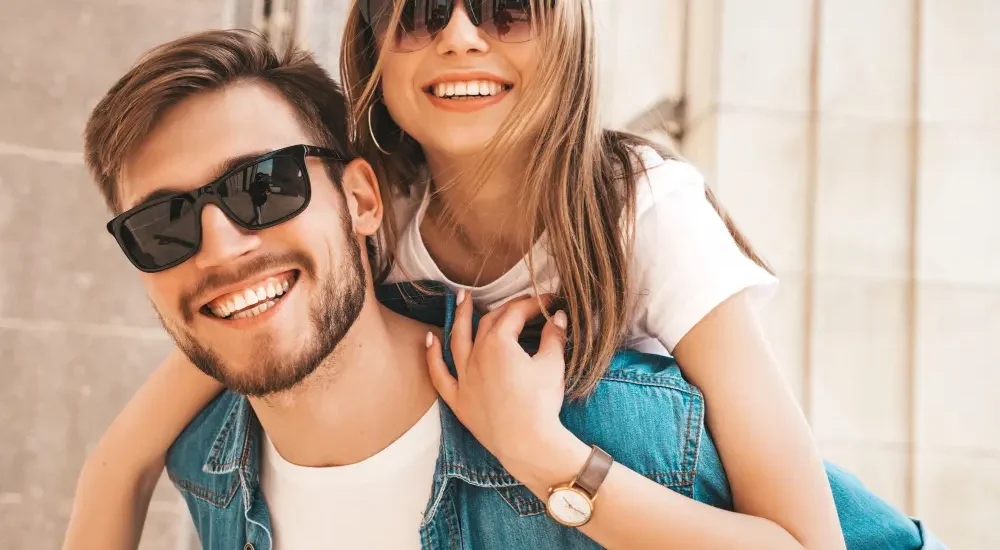 Casal de namorados loiros sorrindo usando óculos de sol com a mulher nas costas do homem o abraçando