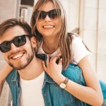 Casal de namorados loiros sorrindo usando óculos de sol com a mulher nas costas do homem o abraçando