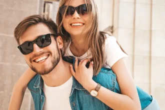 Casal de namorados loiros sorrindo usando óculos de sol com a mulher nas costas do homem o abraçando