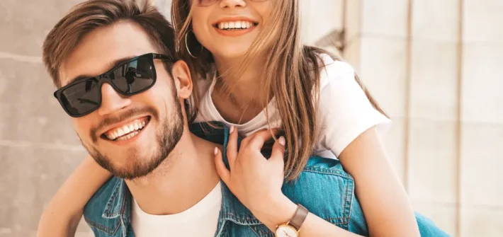 Casal de namorados loiros sorrindo usando óculos de sol com a mulher nas costas do homem o abraçando
