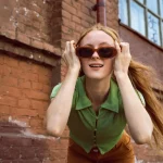 Mulher sorrindo inclinada na direção da câmera segurando os óculos escuros retrô em seu rosto
