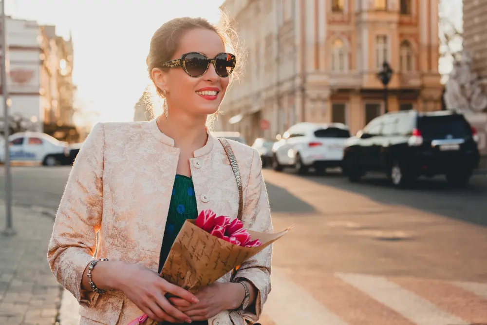 Mulher usando óculos carolina herrera sorrindo segurando buquê de flores rosas na rua com luz natural do sol ao fundo
