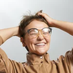 Mulher usando óculos original de grau e sorrindo com as duas mãos no cabelo