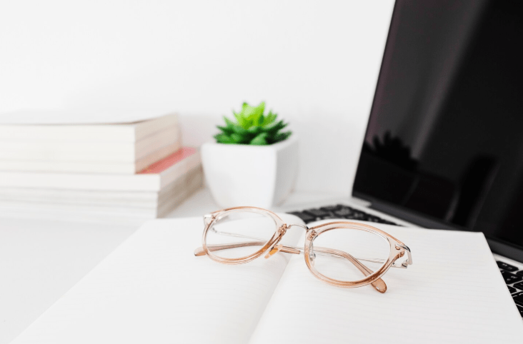 Óculos para trabalho com armação rosada sobre caderno ao lado de notebook em mesa organizada