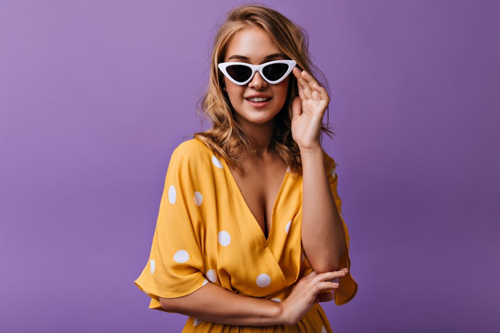 Modelo com blusa amarela de poá exibe óculos Miu Miu original com armação branca estilizada em frente a um fundo roxo.
