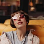 Modelo com cabelo curto exibe modernas armações de óculos de grau feminino de acetato transparente em estilo gatinho sobre sofá amarelo.