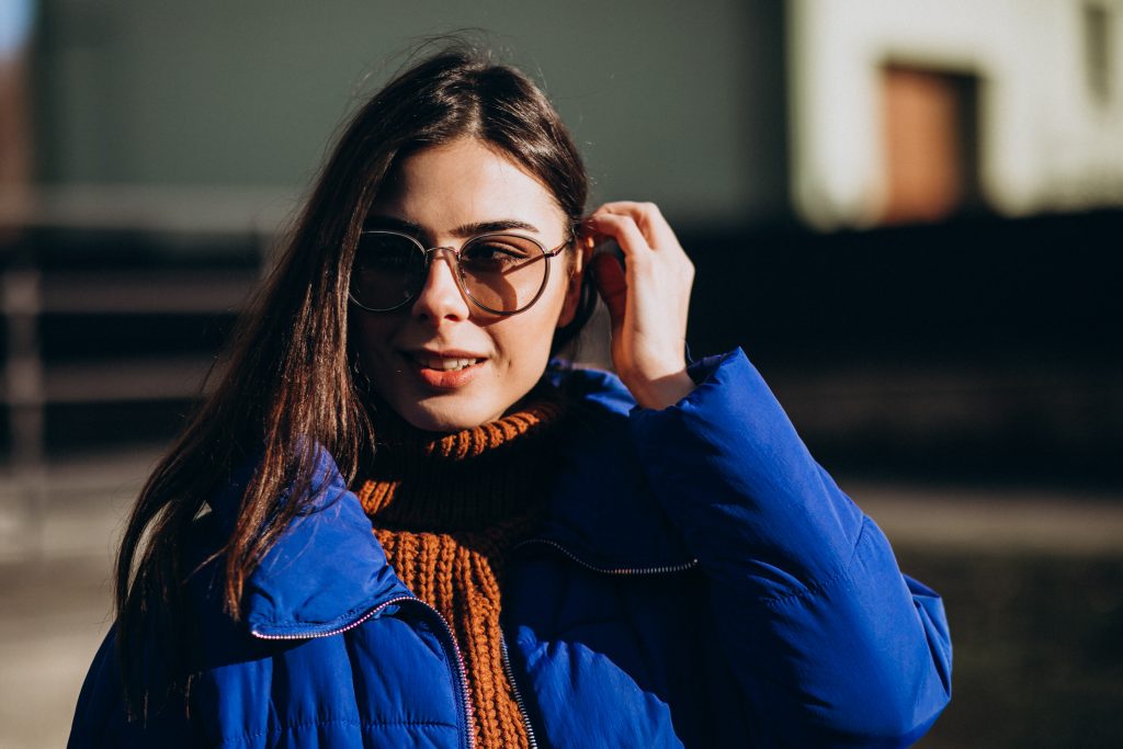 Mulher loira com casaco azul e cachecol marrom usa elegantes armações de óculos de grau feminino metálicas redondas ao ar livre.