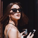 Jovem estilosa segura smartphone e café enquanto destaca seus óculos Tom Ford original de armação gatinho sob a luz do sol.