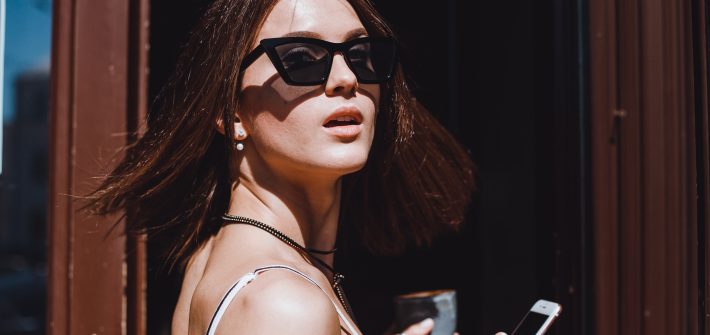 Jovem estilosa segura smartphone e café enquanto destaca seus óculos Tom Ford original de armação gatinho sob a luz do sol.